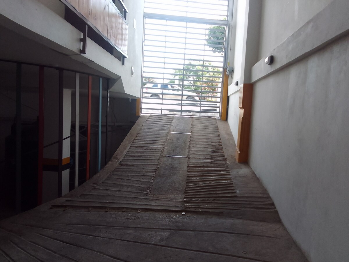 An accessible ramp, made of wooden planks, leads through a bright corridor towards a partially open gate. Natural light enters through the horizontal bars, casting patterns on the floor. The walls are painted in neutral tones, contributing to a clean and functional environment.