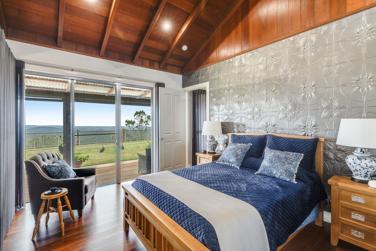 A spacious bedroom features a wooden ceiling and a decorative metallic accent wall. A comfortable bed is dressed in dark blue linens, complemented by patterned pillows. Large sliding doors open to a scenic outdoor view, inviting natural light into the room.
