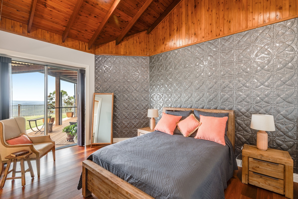 A spacious bedroom features a king-sized bed with a dark quilt and coordinated pillows. Accents of warm wood and a unique textured wall create a cozy ambiance. Natural light flows from glass doors that open to a balcony with scenic views.