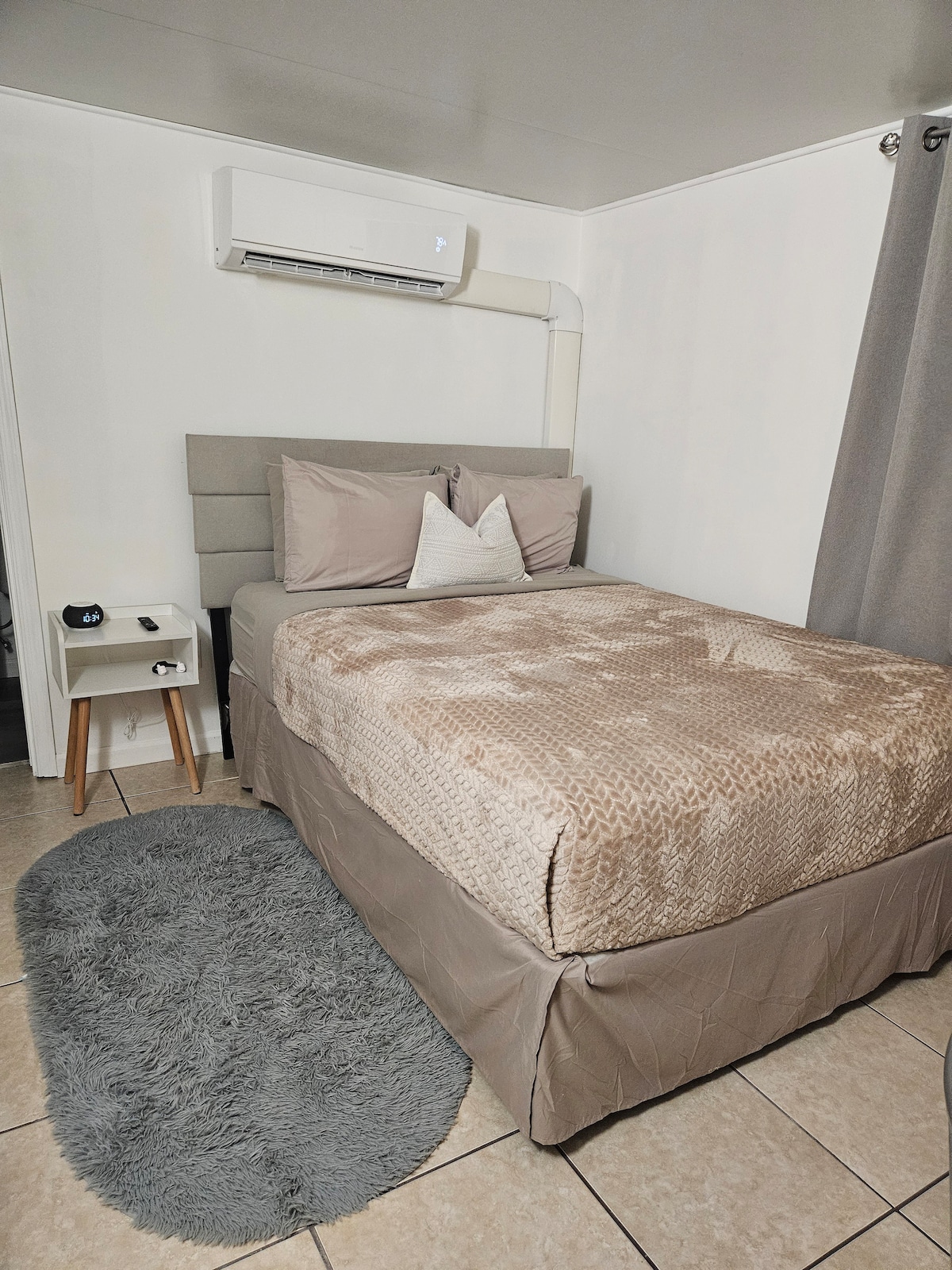 A compact bedroom is shown featuring a cozy bed with a textured blanket and soft pillows. A bedside table with a lamp is positioned next to the bed. Natural light is facilitated by the window, and a simple rug is placed on the tiled floor.