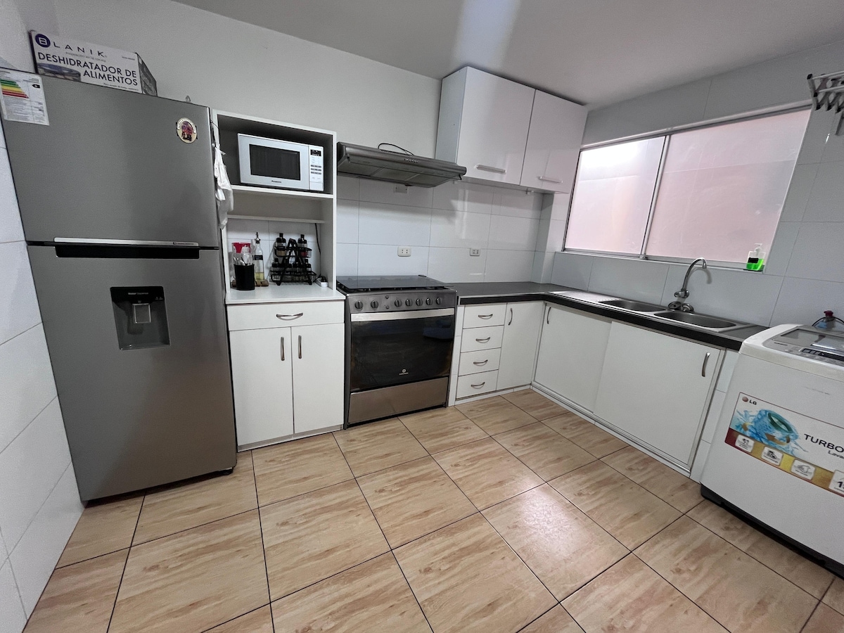 A fully equipped kitchen is featured, showcasing modern appliances including a refrigerator, stove, microwave, and washing machine. Bright natural light enters through a window above the sink, illuminated by light tiles and wooden flooring, enhancing the functional layout of the space.