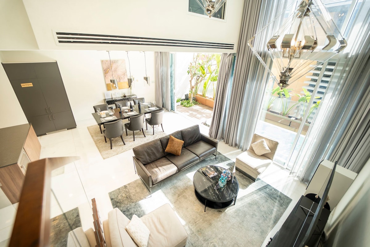 The living area features a modern design with high ceilings, large windows, and an open concept layout. Elegant furnishings include a comfortable sofa and a round coffee table. A dining area with stylish chairs is visible, alongside a private balcony filled with greenery.