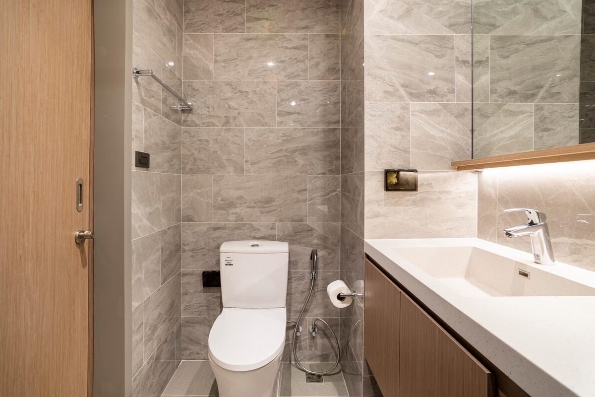 The modern bathroom features textured gray wall tiles and a sleek countertop with a rectangular sink. A wall-mounted mirror reflects the natural light. A toilet with a bidet attachment is positioned beside the sink, and minimalistic wooden cabinetry provides storage.
