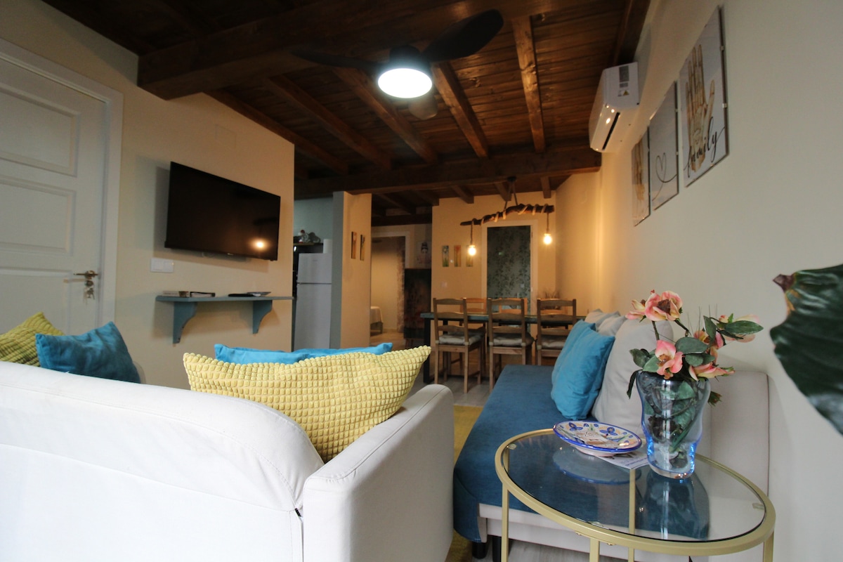 A cozy living area features a white sofa adorned with colorful cushions and a blue chaise. A glass coffee table holds a floral arrangement, and a wall-mounted television is positioned nearby. Exposed wooden beams offer a rustic touch to the space, which leads to a dining area.