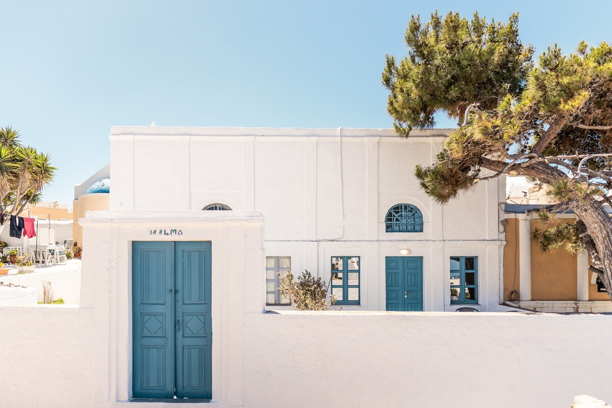 A whitewashed building with a characteristic blue door and window frames is presented against a clear blue sky. Surrounding greenery, including a tall tree, adds an element of nature to the setting. The overall composition reflects a welcoming entrance.