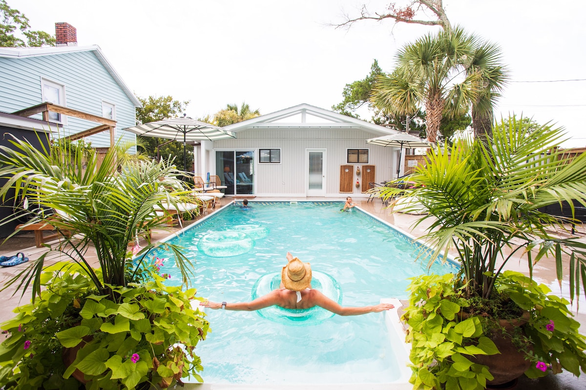 The pool area is framed by lush greenery and potted plants, creating a tropical ambiance. A person is seen floating on an inner tube, while the one-story cottage is visible in the background. Various lounge chairs are positioned around the pool for relaxation.