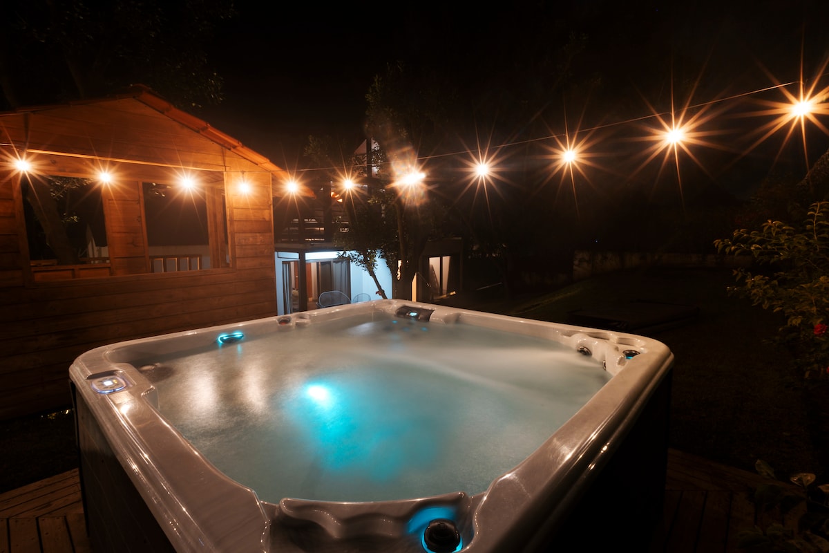 A spacious jacuzzi is illuminated by soft blue lights, surrounded by a wooden deck. String lights hang above, creating a warm ambiance. In the background, a wooden house with large windows is visible, contributing to the serene outdoor setting.