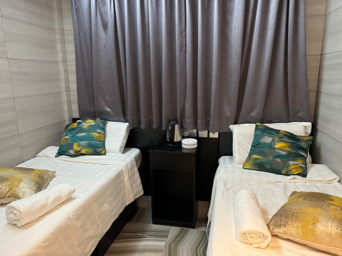 A compact twin bedroom features two single beds with white linens and colorful accent pillows. A small nightstand is positioned between the beds, with a kettle and cups resting on top. Dark curtains cover the window, providing privacy.