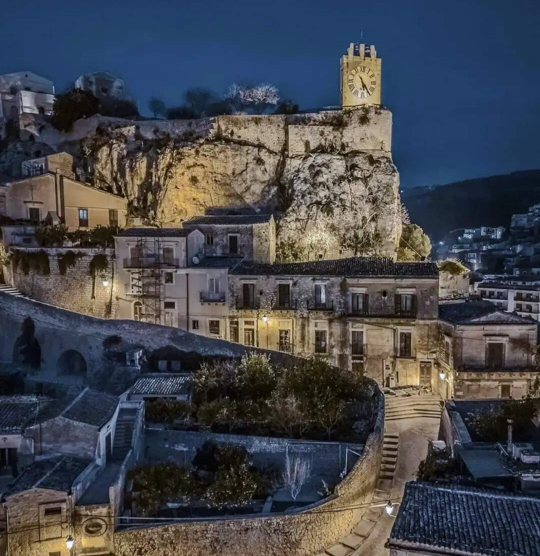 A hillside town is depicted at night, featuring a clock tower atop a rocky elevation. Warm lights illuminate the narrow pathways and historic buildings, creating a serene atmosphere amidst the dark sky. The winding streets reveal a blend of nature and architecture.