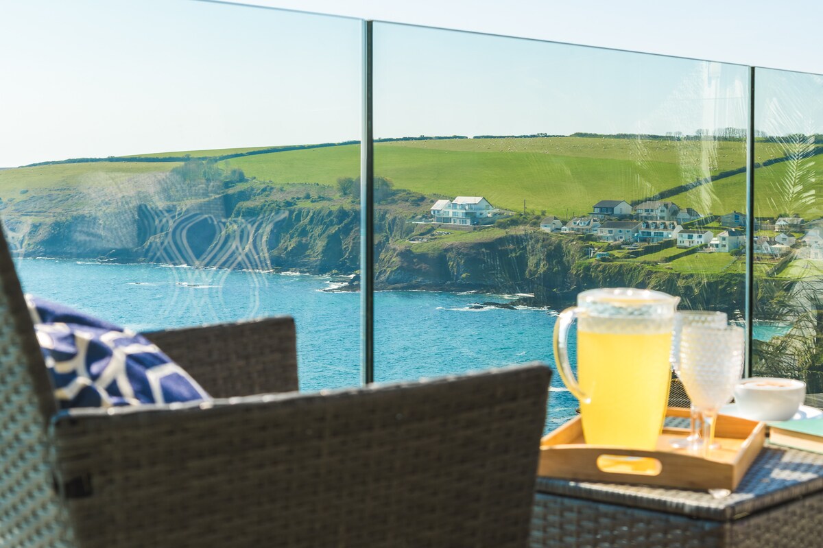 A glass balcony railing offers unobstructed views of the sea and the lush green coastline. A wicker chair features a navy and white cushion, while a tray with a pitcher of lemonade and two glasses is positioned on the table, inviting relaxation.