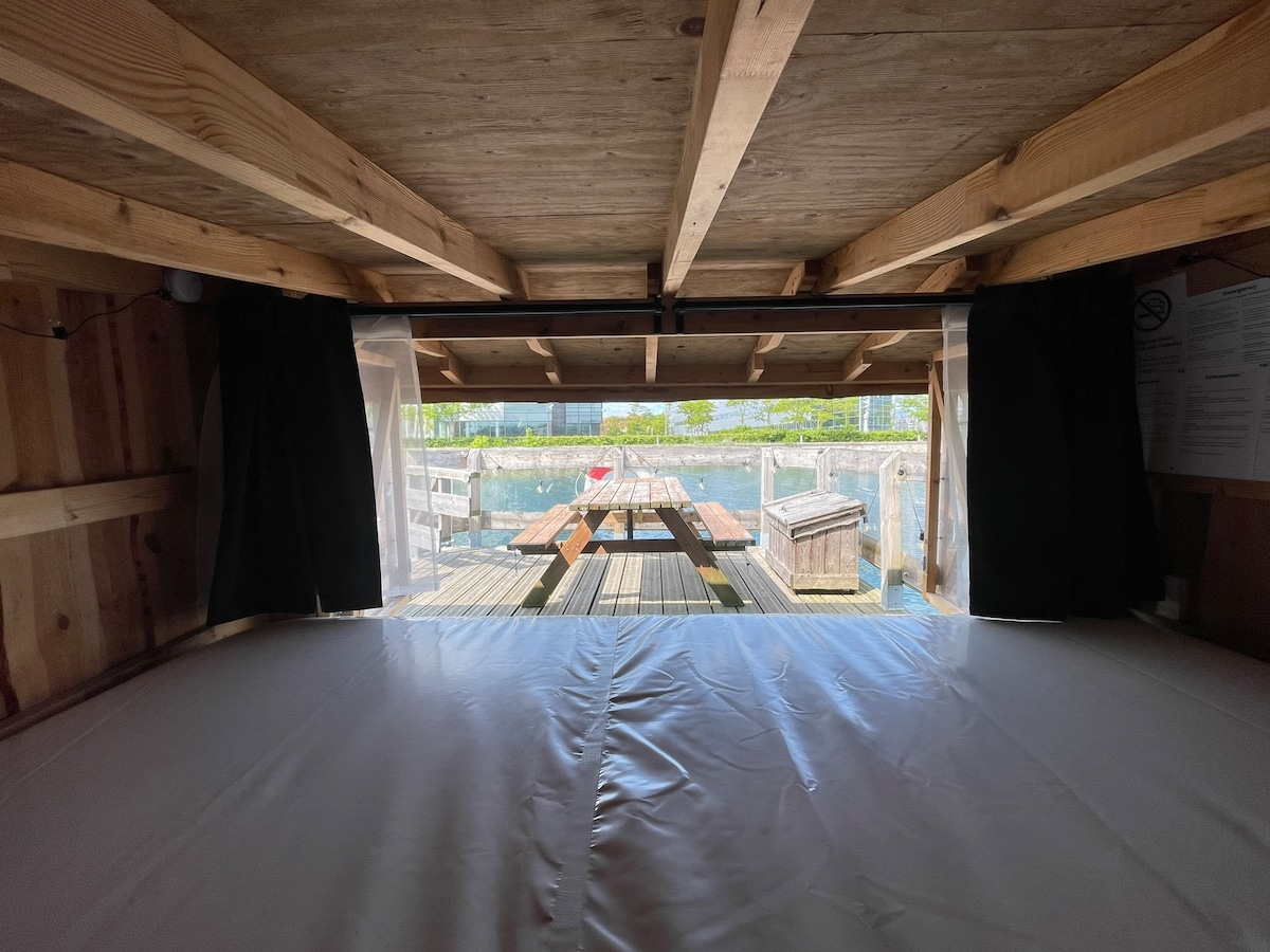 The interior sleeping section reveals a view through large openings framed by dark curtains. A wooden deck with tables and benches is visible just outside, complemented by the surrounding water and greenery, providing a sense of open nature.
