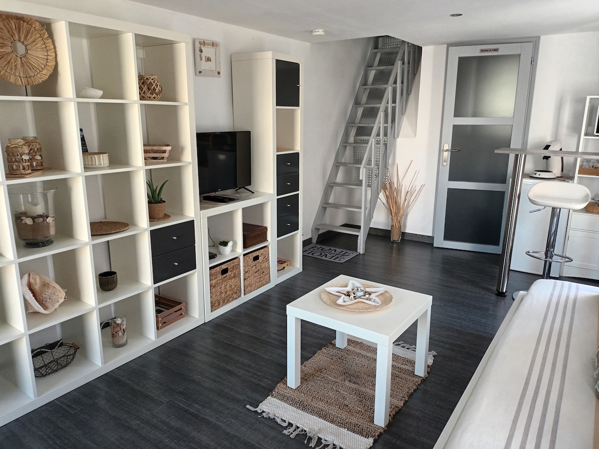 A well-arranged living space features a white shelving unit with decorative items and storage baskets. A flat-screen TV is positioned on a small console. A round table with high chairs stands next to a staircase leading to the upper level, where additional sleeping quarters are located.
