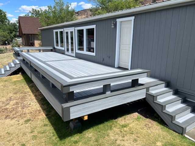 A spacious deck extends from the side of the house, featuring a smooth, gray surface and sturdy railing. Multiple steps lead down to the grassy area, with large windows visible along the side of the home, allowing for ample natural light.