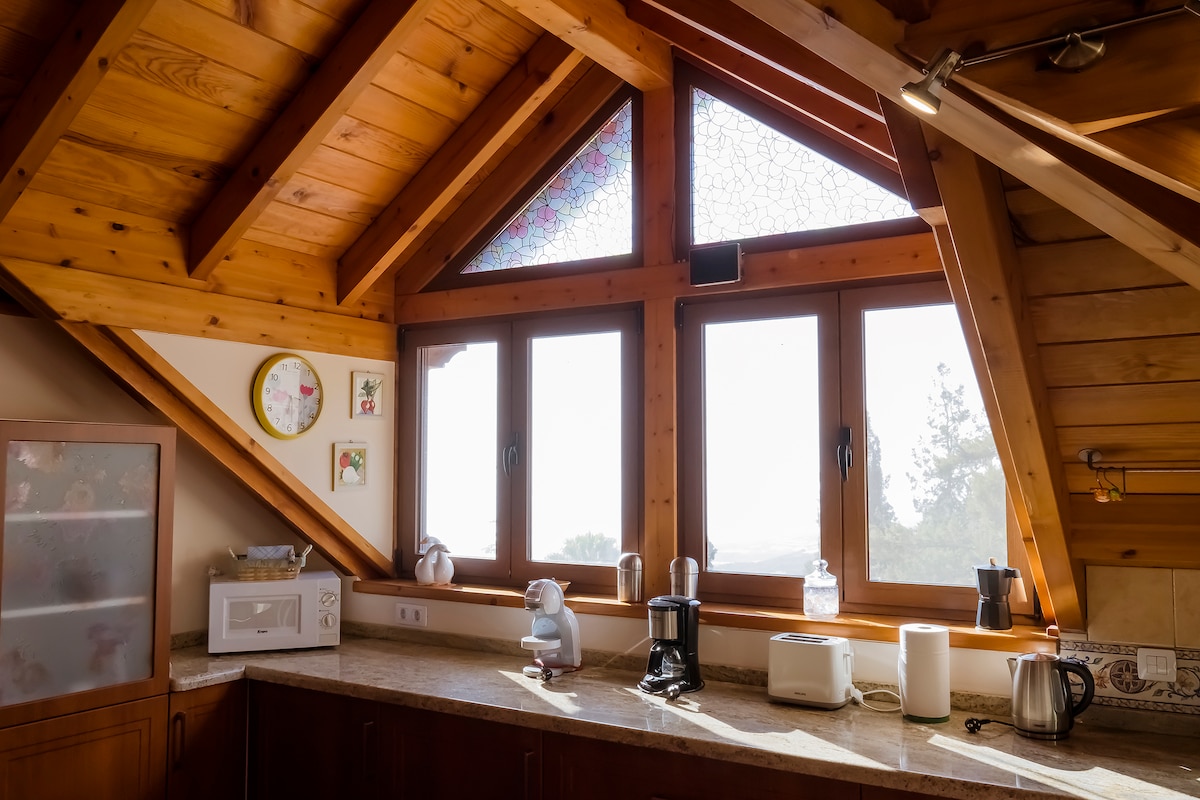A well-equipped kitchen features wooden beams and large windows with stained glass accents, allowing natural light to enter the space. Kitchen appliances, including a microwave, coffee maker, and kettle, are arranged on the countertop, providing functionality within a warm, inviting atmosphere.