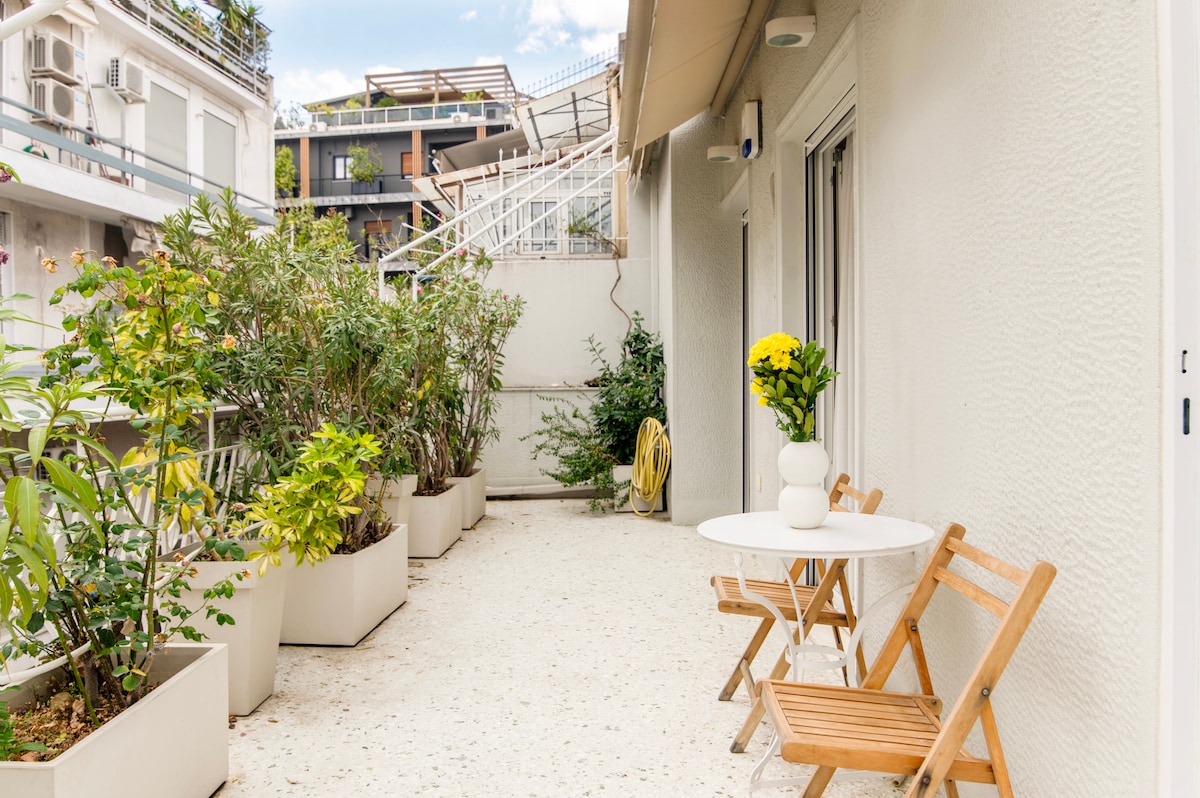 A private terrace area is showcased, featuring a light-colored surface and several potted plants that provide a touch of greenery. A small round table is paired with two wooden chairs, offering a space for relaxation outdoors. Bright yellow flowers are displayed in a white vase.