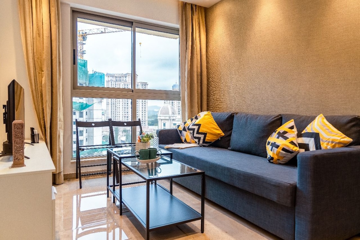 A modern seating area is presented, featuring a spacious grey sofa adorned with yellow and black accent pillows. A glass coffee table sits in front, complemented by a small decorative centerpiece. Large windows provide natural light and offer views of the city skyline.
