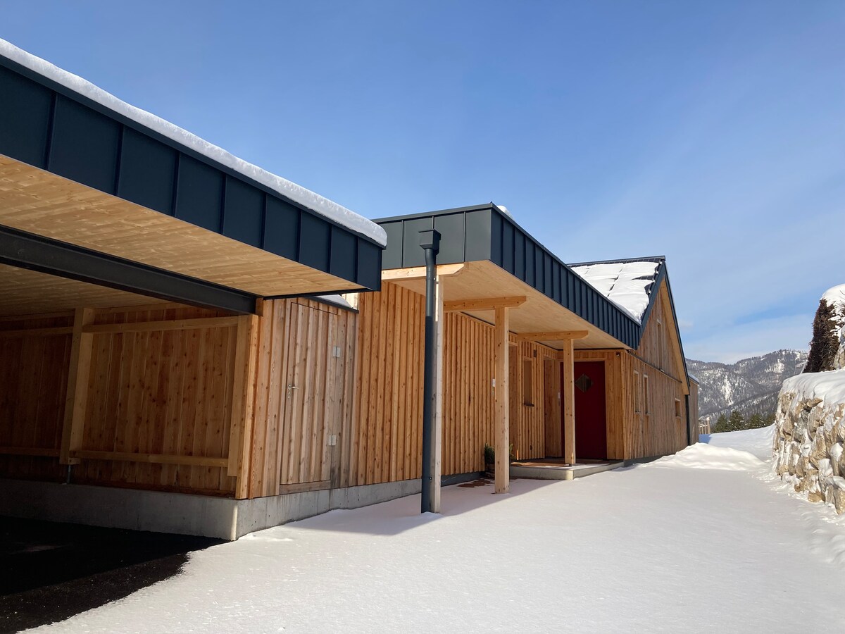 A modern wooden structure is presented in a snowy landscape, featuring a carport on the left. The entrance is marked by a bright red door, complemented by natural wood paneling and a sloped roof. Mountains and a clear sky are visible in the background.