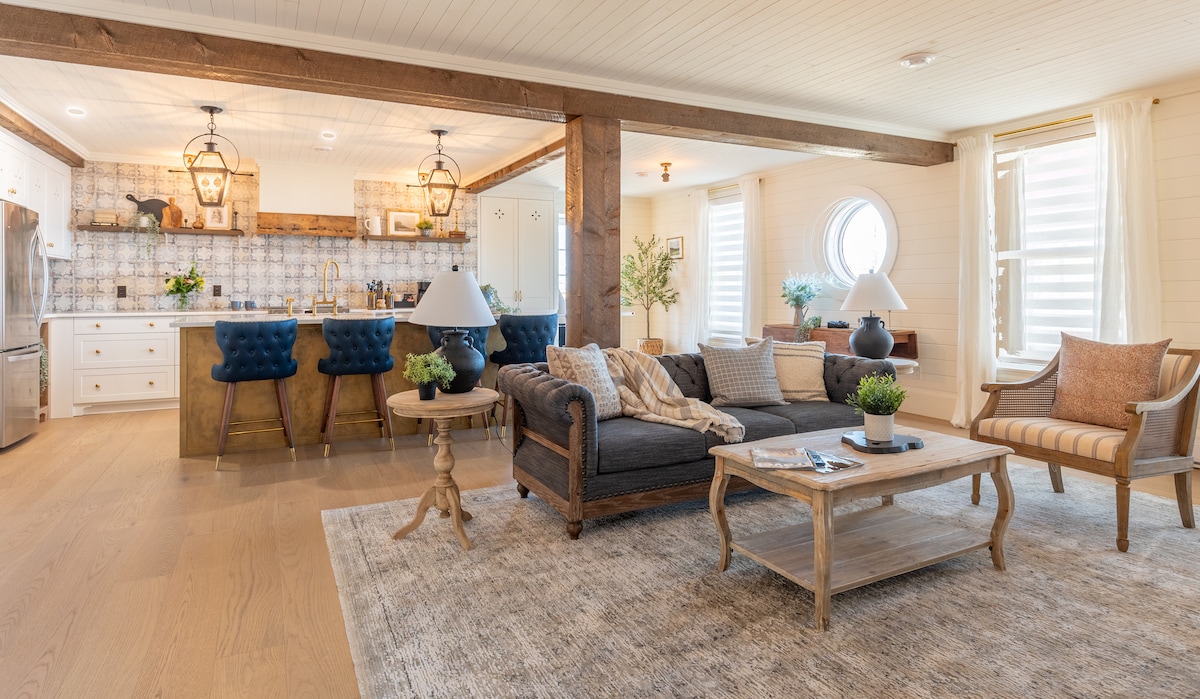 An open-concept living space features a blend of light wood and brick accents. A dark sofa and a pair of striped chairs are arranged around a wooden coffee table. A kitchen island with blue barstools is visible in the background, along with a porthole window allowing natural light.