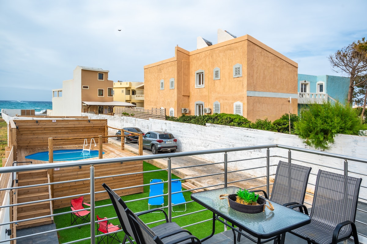 A spacious outdoor terrace offers a dining table surrounded by chairs, with a pool visible nearby. The backdrop includes a light-colored building and greenery, with the sea visible in the distance. Sunlight brightens the scene, creating an inviting space for relaxation.