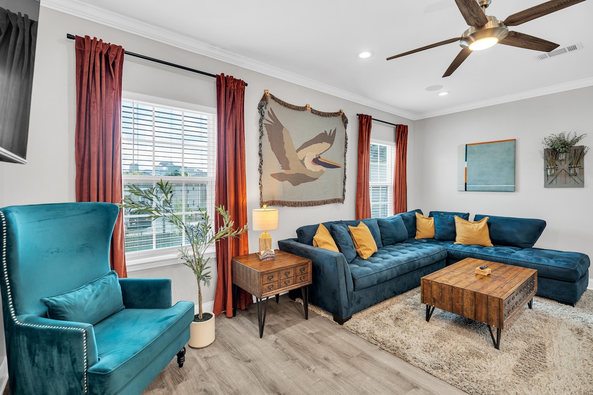 A spacious living area is presented, featuring a large blue sectional sofa accented with yellow pillows. A wooden coffee table sits in the center, and a wall-mounted TV is positioned nearby. Floor-to-ceiling windows are adorned with red curtains, complemented by a decorative tapestry and indoor plants.