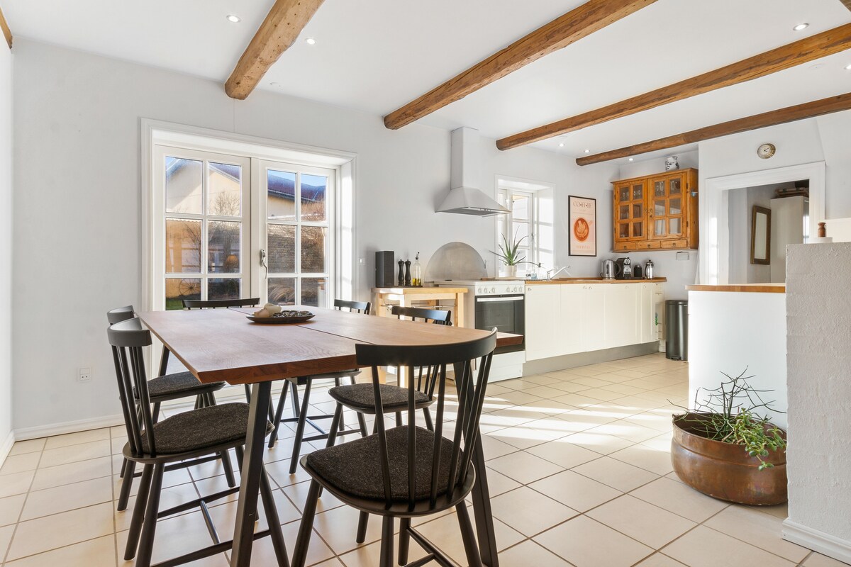 A light-filled kitchen and dining area features a wooden table surrounded by six black chairs. Natural light streams through large windows, highlighting wooden beams above. The kitchen is equipped with modern appliances and has a neatly organized layout, contributing to a functional cooking space.