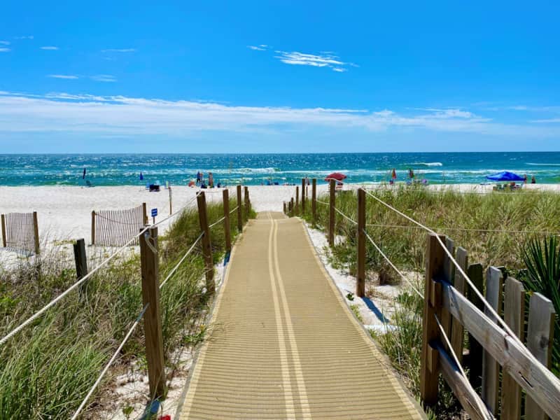 A wooden boardwalk leads through sandy dunes, offering a clear view of the beach and ocean. Umbrellas are visible on the beach, while gentle waves roll in under a bright blue sky with a few scattered clouds.
