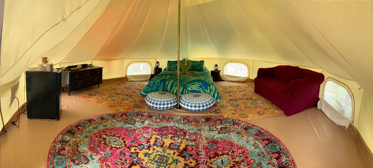 An inviting interior space features a large bed adorned with tropical-themed linens, surrounded by a vibrant patterned area rug. A plush loveseat and decorative poufs offer additional seating. Soft lighting is provided by lamps on either side of the bed, enhancing the cozy atmosphere.