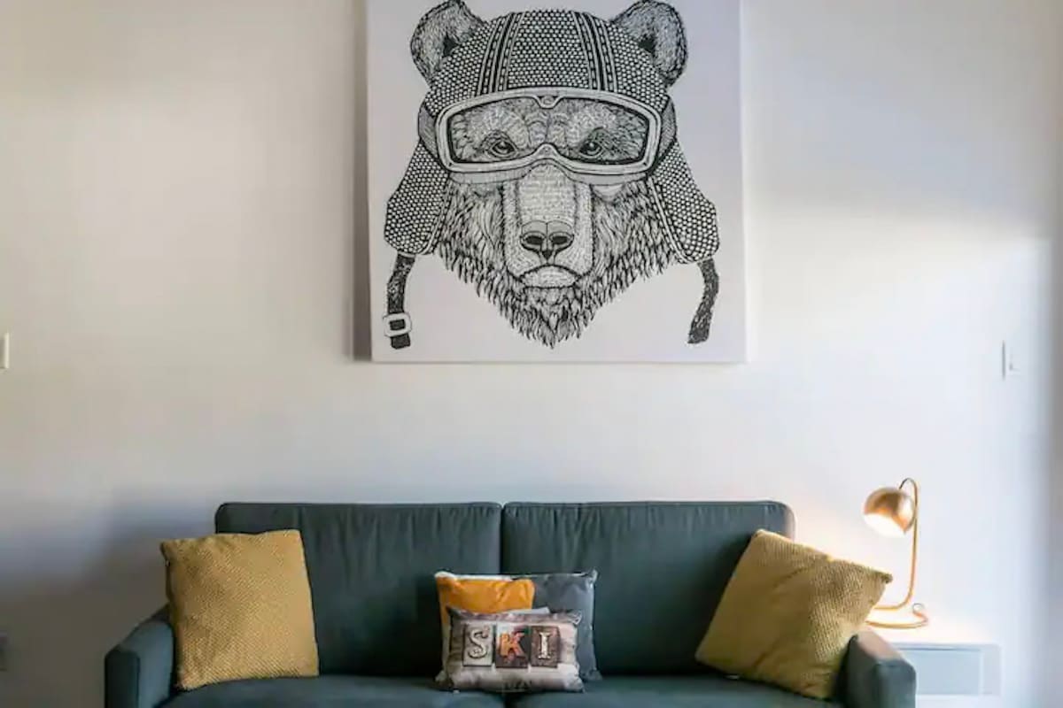 A modern living area features a comfortable green sofa with two yellow accent pillows. Above the sofa, a large artwork of a bear wearing skiing goggles adds a playful touch to the space. A stylish lamp stands in the corner, completing the cozy ambiance.