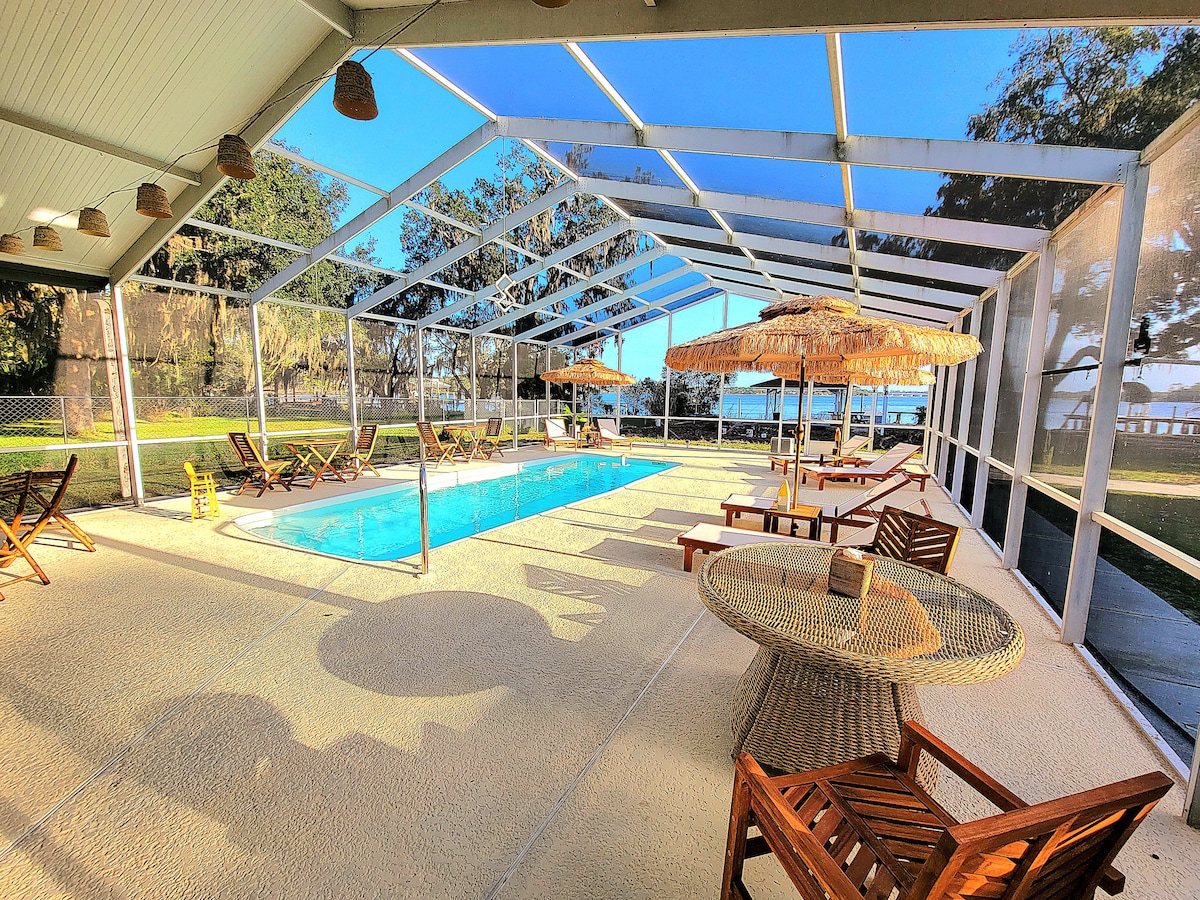 A screened-in pool area is displayed, featuring a refreshing blue pool under clear skies. Teak lounge chairs and a circular table are arranged around the pool, while thatched umbrellas provide shade. A scenic view of the surrounding landscape is visible through the screens.