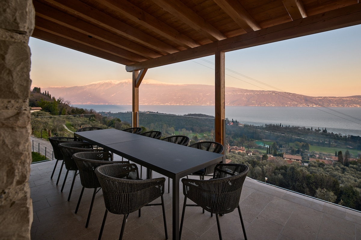 An outdoor terrace features a long dining table surrounded by six stylish chairs, all set against a backdrop of scenic lake views and distant mountains. The space is sheltered by a wooden roof, offering an inviting area for outdoor gatherings.