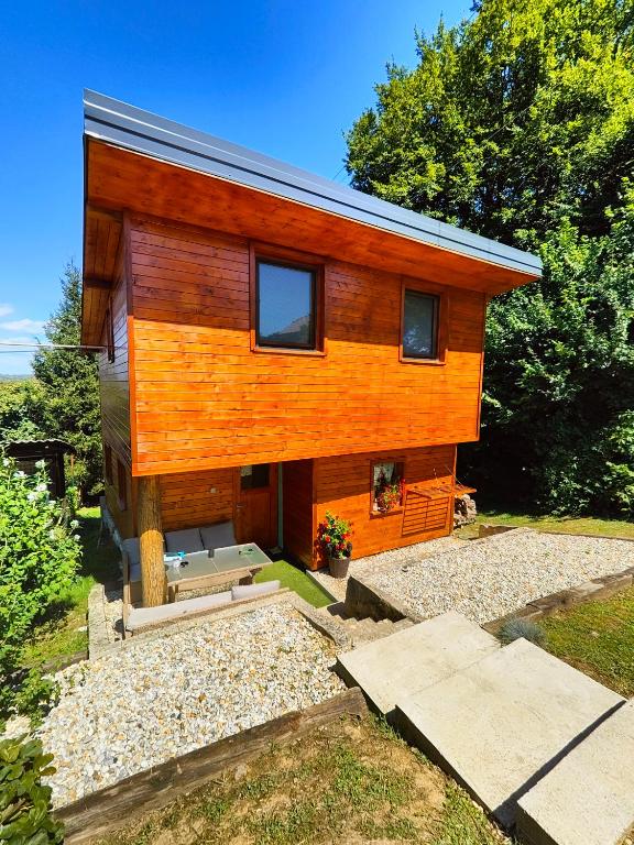 A modern wooden house with a distinctive orange façade is surrounded by greenery. Two large windows allow natural light to flow in. A pathway made of stone leads up to the entrance, while flowering plants add a touch of color to the setting.