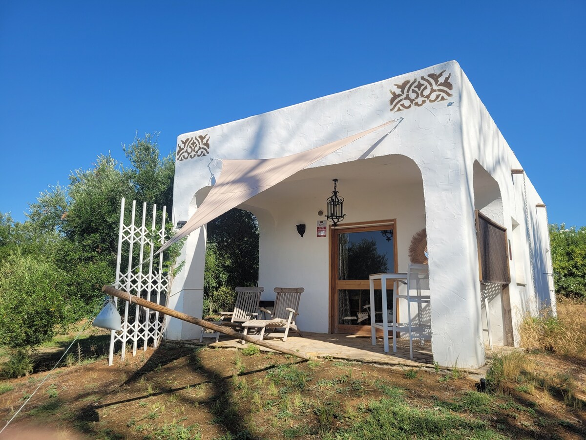 The bungalow's exterior is characterized by a white façade with decorative elements above the entrance. A light-colored awning provides shade over the entry area, where wooden chairs are positioned beside a small table. Lush greenery surrounds the property, enhancing the serene setting.