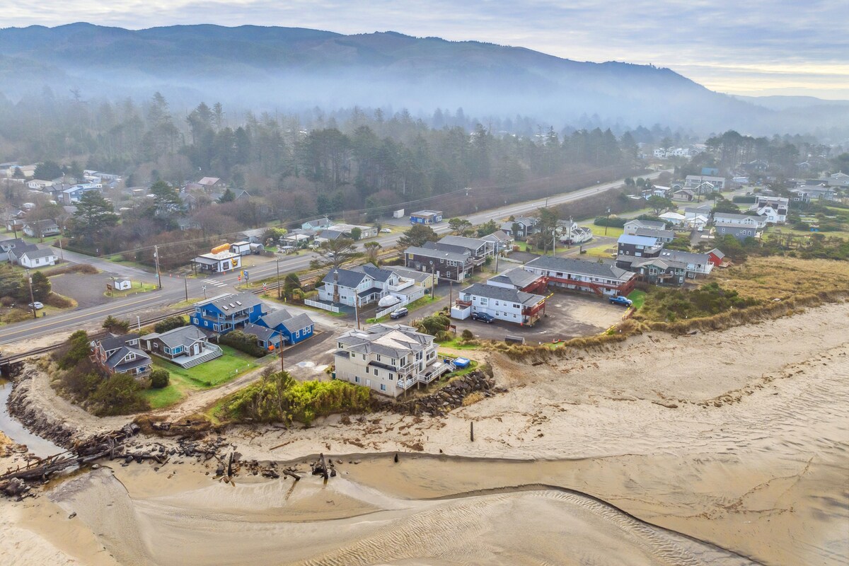 An aerial view captures the coastal landscape, featuring a blend of residential homes and open sandy spaces. The shoreline is bordered by gentle dunes, while the distant mountains rise softly in the background. This image emphasizes the proximity to the beach and surrounding area.