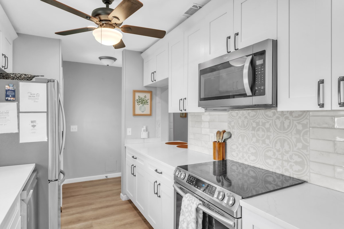A modern kitchen features white cabinetry and a dark countertop accentuated by stylish backsplash tiles. Stainless steel appliances, including a microwave and refrigerator, are visible. A ceiling fan adds functionality, while natural light illuminates the space, enhancing its bright and open layout.