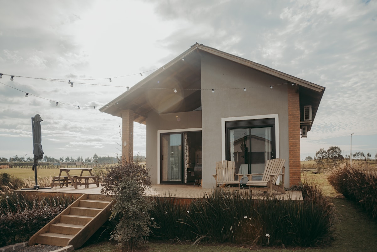 A welcoming exterior of a single-story chalet is visible, featuring large glass doors and wooden decking. Two adirondack chairs are placed on the porch, while a picnic table is seen nearby. Soft landscape lighting is strung along the eaves, enhancing the outdoor space.