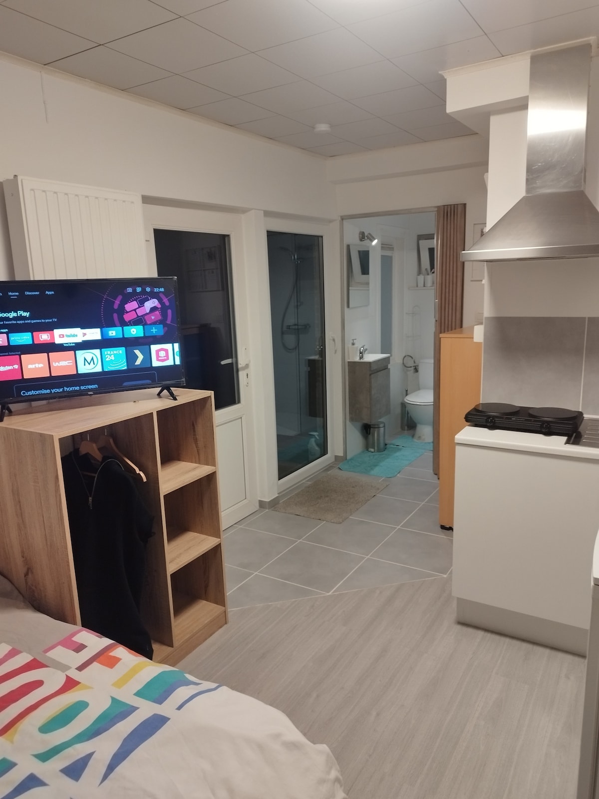 A compact living space features a television mounted on a wooden shelf, with a visible closet for clothing. A neat bathroom area is accessible through a glass door, while a well-equipped kitchenette showcases simple cabinetry and appliances against a tile backdrop.
