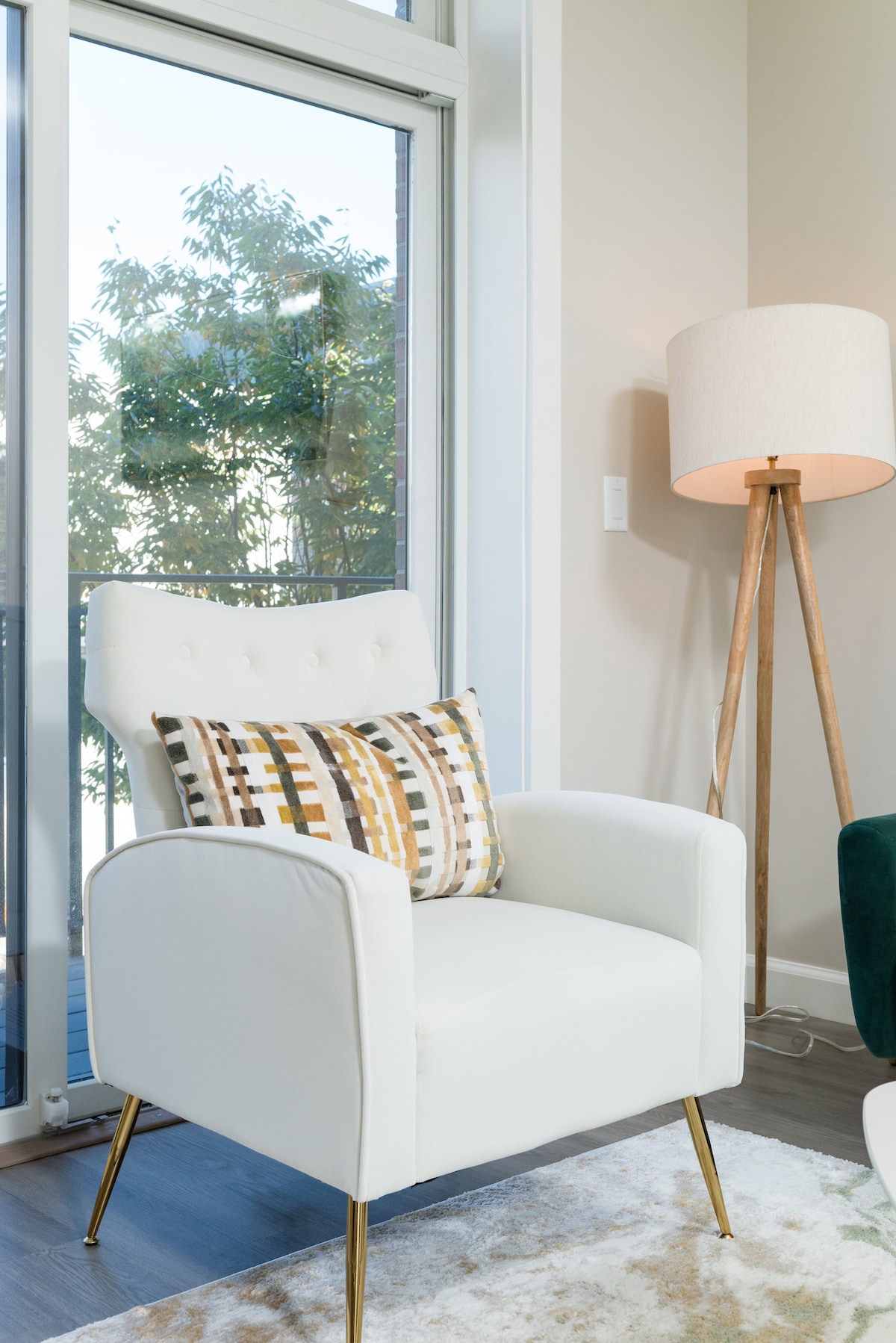 A modern white armchair with a decorative pillow is positioned near a large window, allowing natural light to fill the space. A tall, simple lamp stands nearby, adding a touch of warmth to the cozy corner.