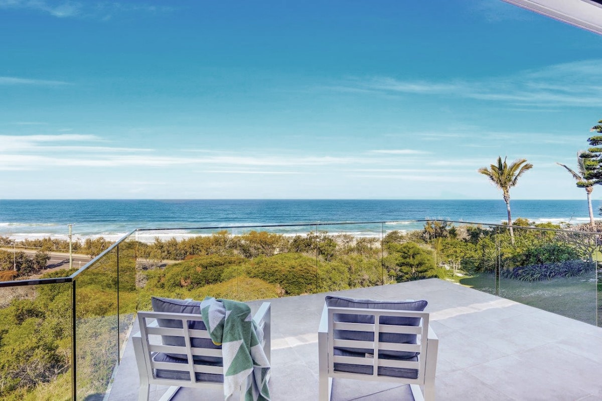 A serene outdoor balcony is showcased, featuring two white lounge chairs facing the ocean. A soft green throw is draped over one chair. The expansive view reveals the blue ocean, sandy beach, and coastal greenery under a clear sky.