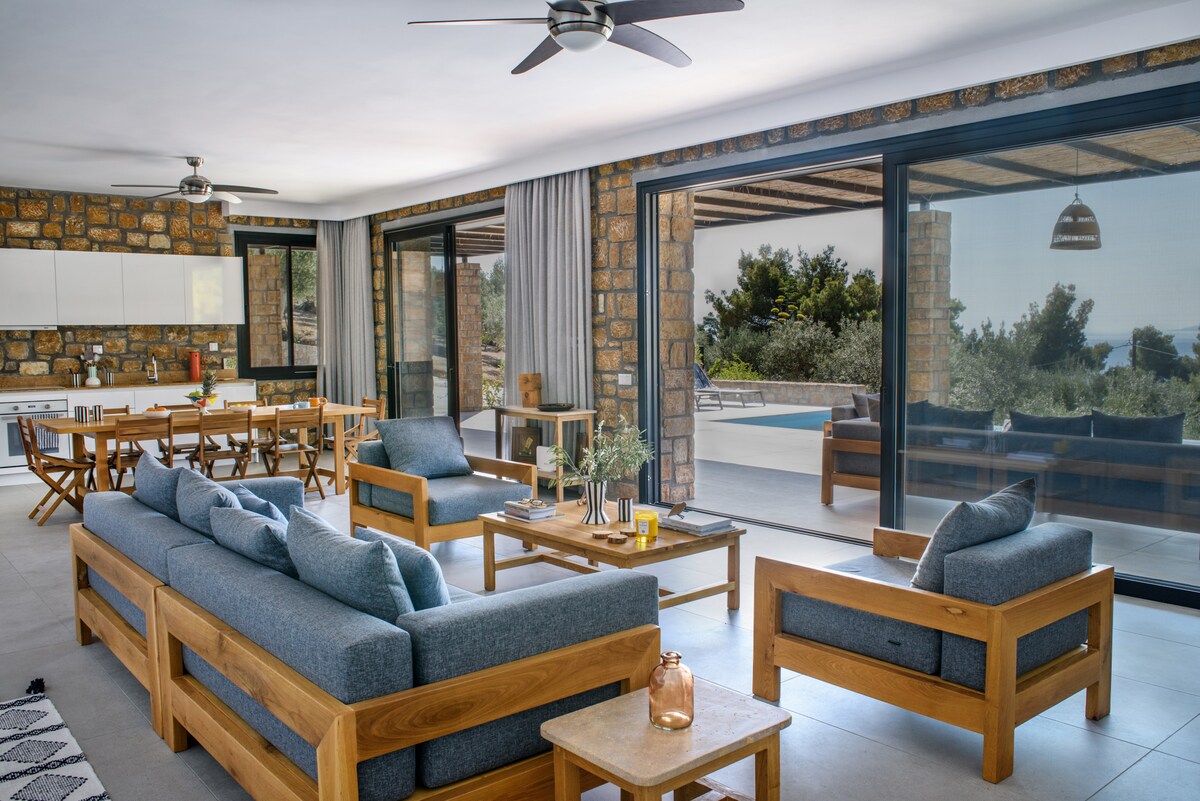 An open-plan living and dining area features handcrafted oak furniture and large glass doors that welcome natural light. The seating arrangement includes comfortable sofas with decorative cushions, accompanied by a wooden coffee table. Views of the outdoor pool and garden are visible through the expansive doors.