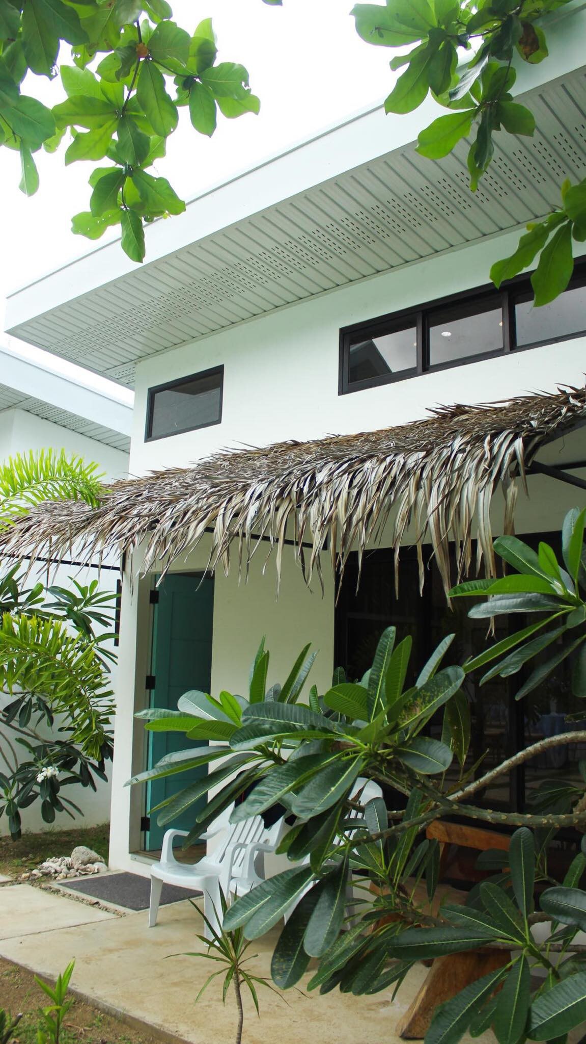 A charming bungalow is framed by lush greenery and tropical plants. A thatched roof offers shade to a small patio area, which features white plastic chairs. Light filters through large windows, and a serene outdoor setting is visible.