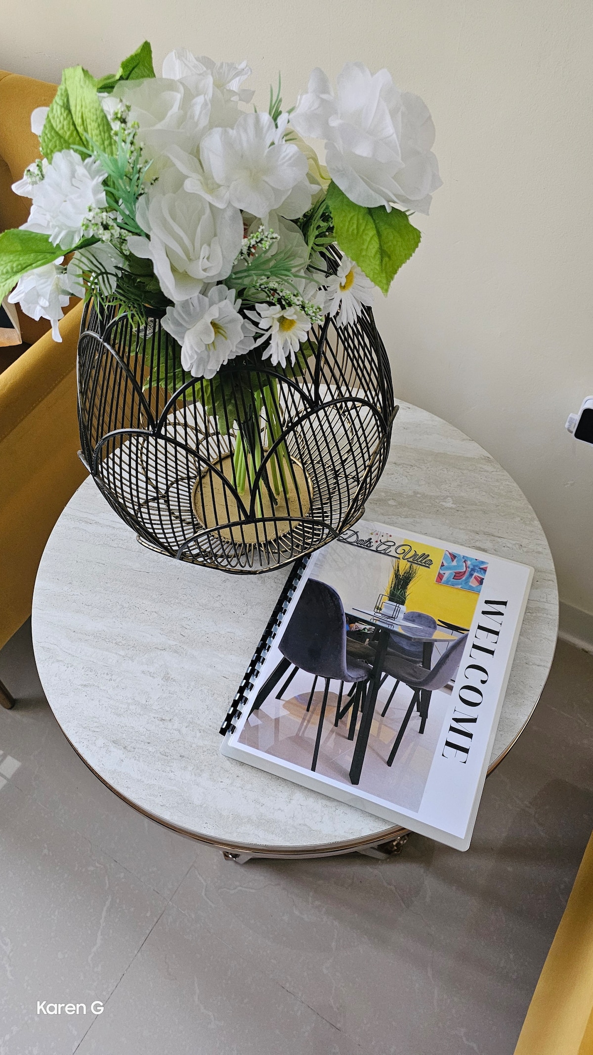 A round table is adorned with a decorative wire vase filled with artificial flowers, providing a touch of greenery. A welcome booklet, featuring a stylish design, is placed beside the vase, inviting guests to explore the home.