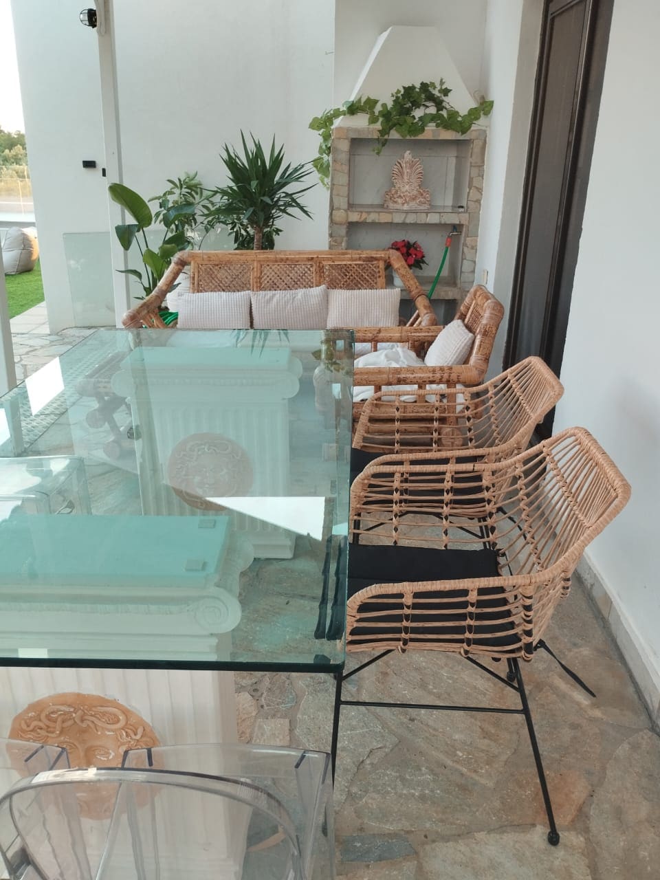A sunny outdoor seating area is showcased, featuring a glass table surrounded by woven chairs. A cushioned sofa is positioned nearby, along with potted plants that add a touch of greenery to the space.
