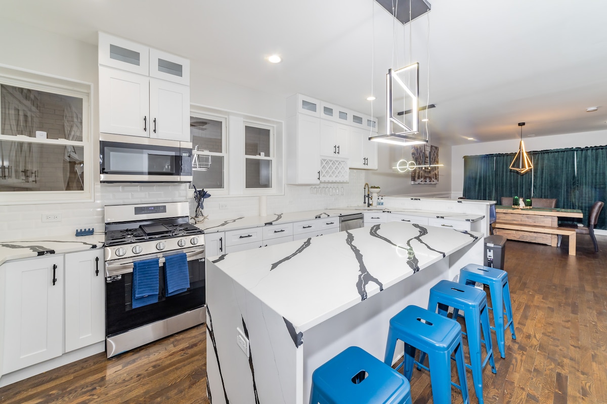 A modern kitchen features white cabinets and an expansive marble countertop with striking vein patterns. Stainless steel appliances, including a gas stove, are visible. Four blue metal stools line the counter. Natural light floods the room, highlighting an adjacent dining area with a wooden table.