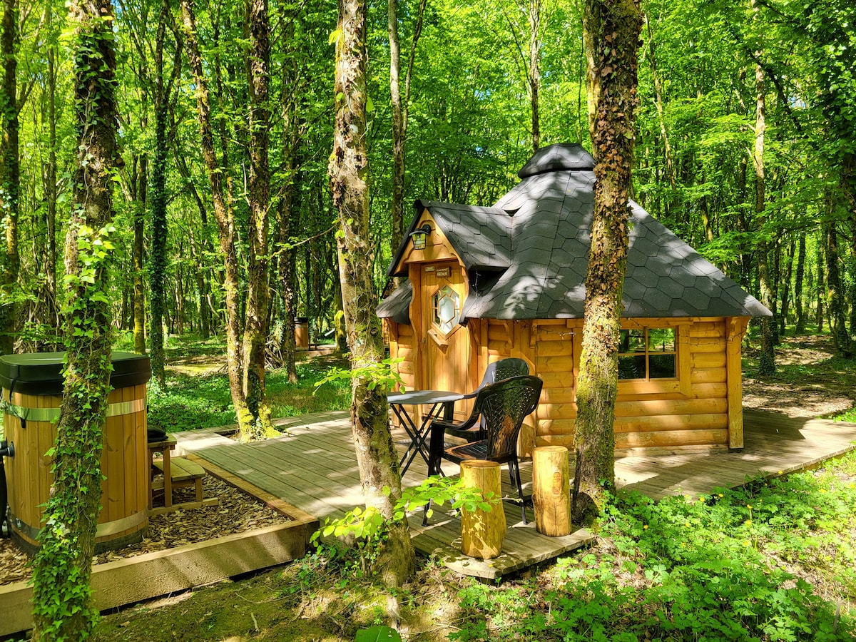 A cozy wooden cabin is nestled among lush green trees, featuring a distinctive round shape and a sloped roof. A small wooden deck with a table and chair is positioned outside the cabin, while a private Nordic bath is visible nearby.