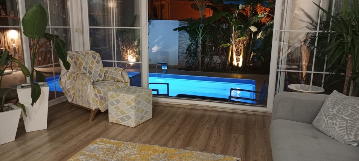 A seating area is visible with a patterned armchair and ottoman beside a spacious light-colored sofa. A plush rug is placed on the wooden flooring. Large windows showcase a swimming pool and landscaped greenery illuminated by outdoor lighting.