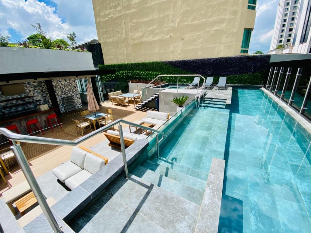 A modern pool area features a long, crystal-clear swimming pool, with steps leading in from a sun deck. Seating areas with wooden furniture are positioned underneath shade structures, and lush greenery lines the perimeter, creating a relaxing environment.