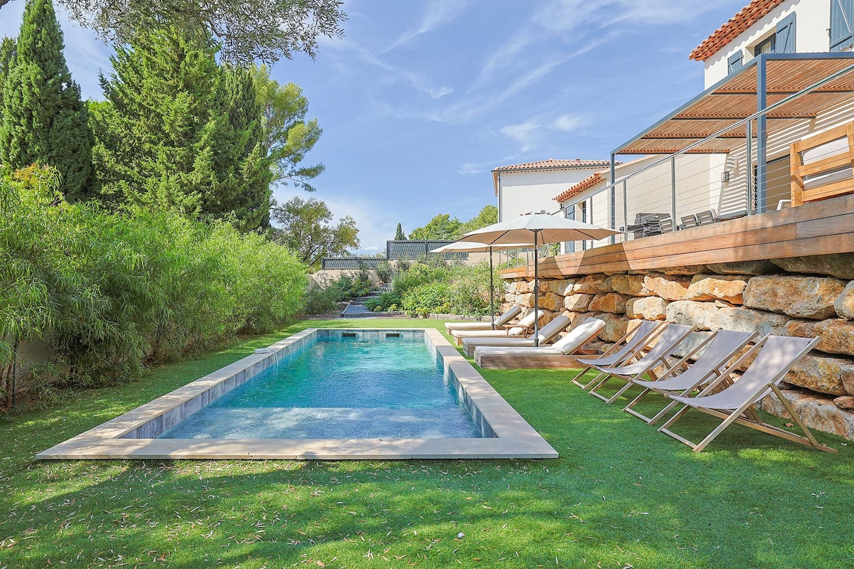 A serene outdoor area features a rectangular, heated salt-water pool surrounded by green grass. Six lounge chairs are positioned on the side, facing the water. A shaded area with an umbrella provides respite from the sun, accentuating the tranquil ambiance.