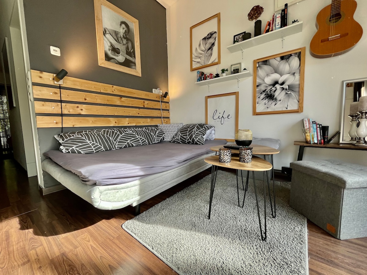 A cozy sleeping area is presented, featuring a spacious bed with patterned bedding against a wood-accented wall. Decorative shelves hold various items, while framed artwork decorates the surrounding walls. A small coffee table and pouf complement the space, with soft light illuminating the floor.