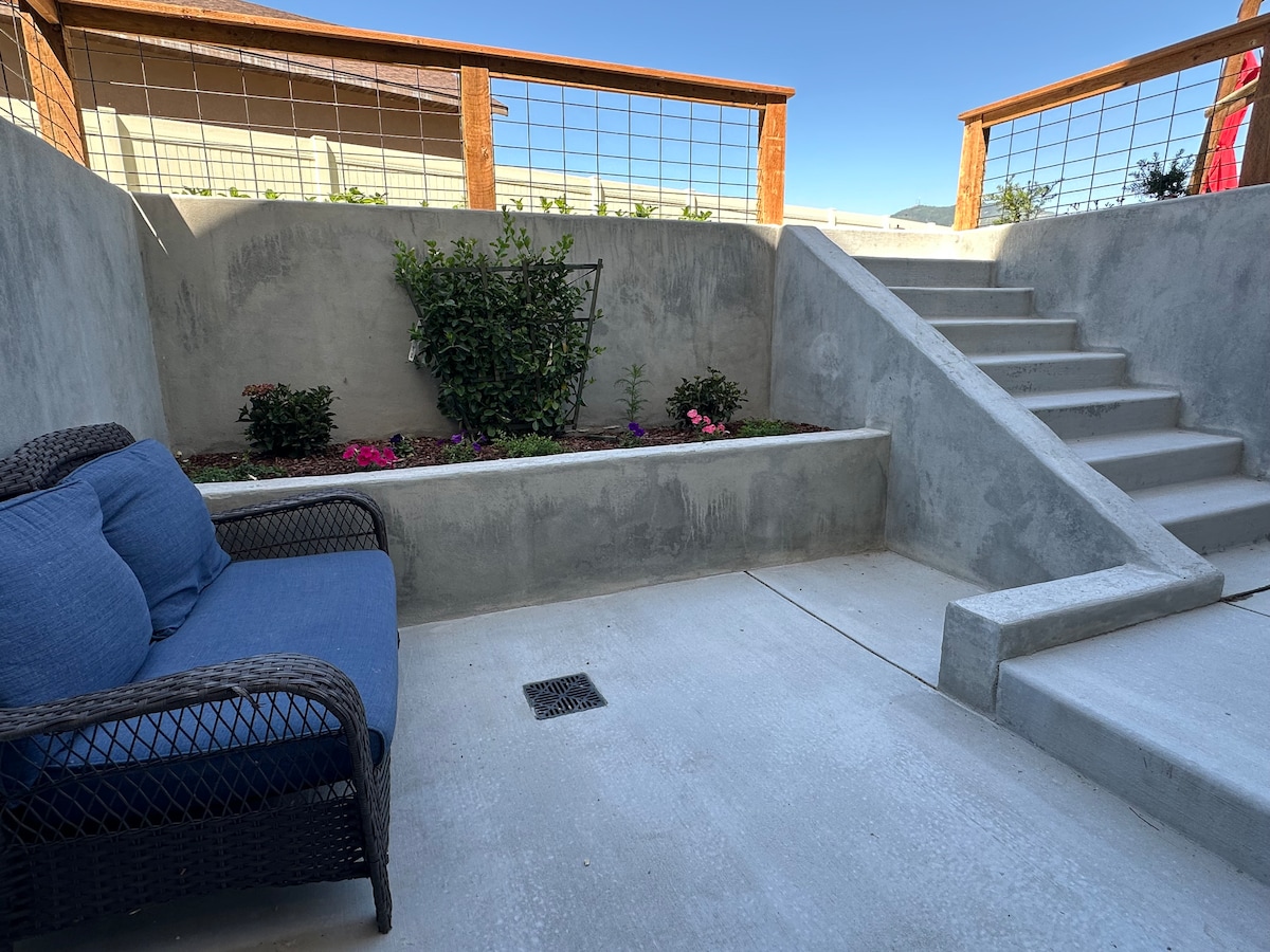 An outdoor seating area features a comfortable blue sofa set against a concrete wall. Steps lead to an upper level, while planted shrubs and flowers enhance the area. A wooden railing adds a natural touch to the space.