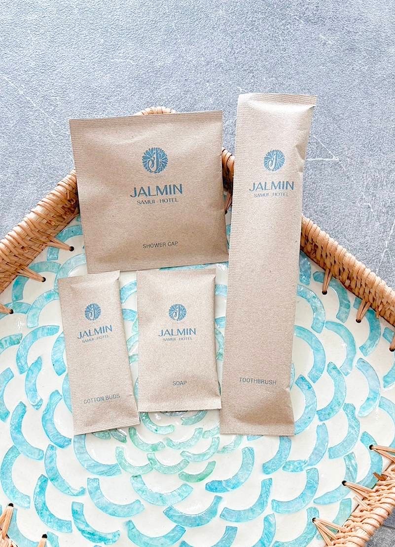 A neatly arranged display of JALMIN Hotel amenities is shown on a decorative tray. The items include a shower cap, cotton brush, soap, and toothbrush, all packaged in minimalistic, eco-friendly wrappers featuring the hotel's logo.
